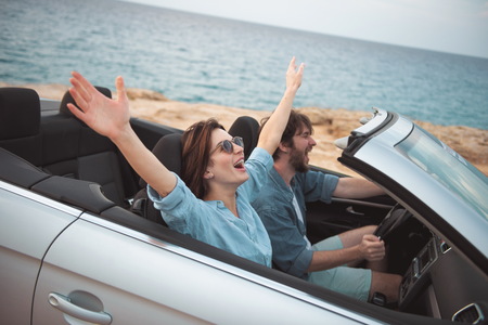 Full of joy. Overjoyed young romantic tourists couple is riding by luxury cabriolet along road with seascape in background. Woman is sitting with raised hands while enjoying summer vacationの写真素材