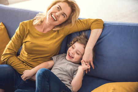 Cheerful parent is enjoying spending great time with kid at home. They are sitting side by side and...