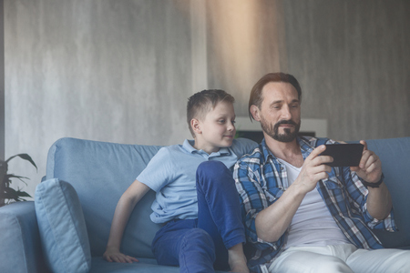 Portrait of cheerful father and satisfied child looking at mobile while situating on sofa. Happy parent and son spending time together conceptの写真素材