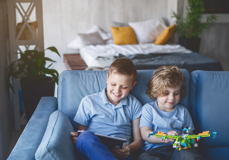 Portrait of happy kid playing toy while sitting near glad brother. He using digital device