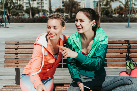 Cheerful female friends are gossiping while sitting on bench in the park. Girl is pointing finger...
