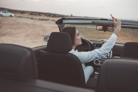 Cheerful young woman is making selfie during her summer trip. She is sitting at steering wheel in her vehicle and smiling. Lady is looking at cellphone camera with joyの写真素材