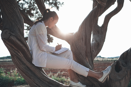 Inspired by nature. Peaceful young woman is sitting on tree and writing a poem into notebookの写真素材