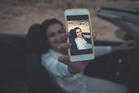 Joyful female tourist is traveling by car in picturesque places. She is making selfie on mobile phone while sitting at steering wheel and smiling. Focus on gadget screenの写真素材