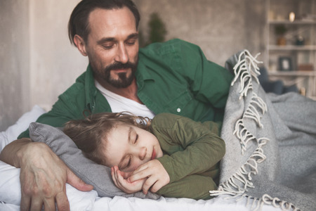 Portrait of smiling dad looking at calm sleeping son in apartmentの写真素材