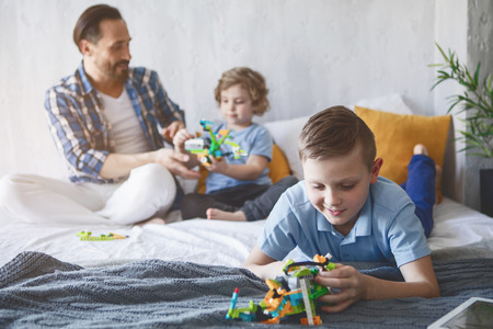 Outgoing elder brother playing with toy alone while little kid spending time with glad dad. They sitting on sofaの写真素材
