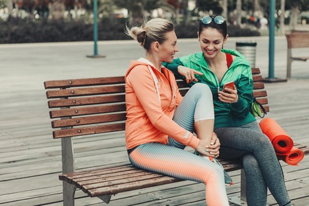 Happy two girls are talking and laughing while sitting on bench outdoor. Lady is showing smartphone to her friend. They are wearing sportswearの写真素材