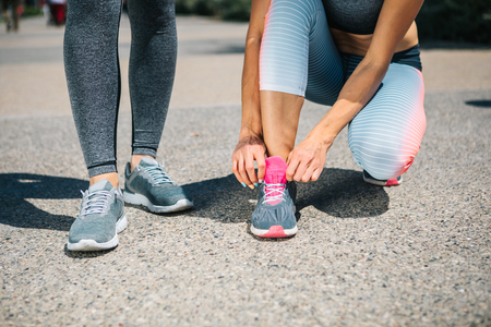 Close up of female feet. Young woman is kneeling and typing laces on sneaker. Her friend is standing on the road and waitingの写真素材