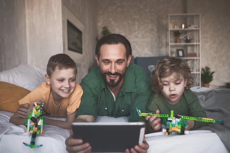 Portrait of outgoing brothers and satisfied dad looking at electronic tablet while relaxing on bedの写真素材