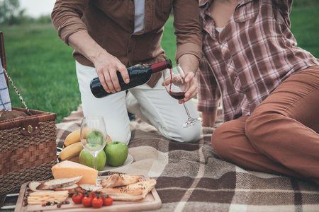 Close up of man arm pouring red wine from bottle into glass. Woman is sitting near husband on the blanket and relaxing. Romantic picnic on grass field concept の写真素材