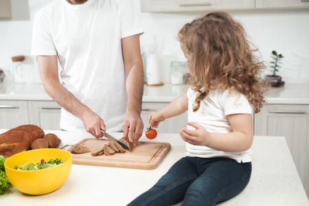 Pretty girl is helping her father to cook lunch. Man is cutting bread into pieces while standing at table neat his child in kitchen  の写真素材