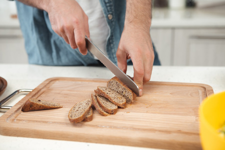 Close up of male hands cutting fresh bread with crust into thin pieces on wooden board. Man is...