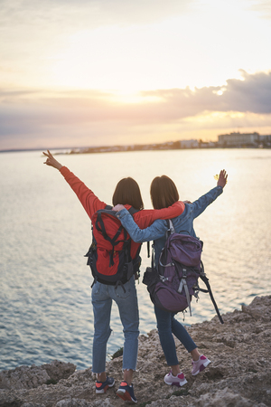Traveling around the world. Excited two young women are watching sunset at the sea together. They are embracing and raising hands up. Real friendship conceptの写真素材