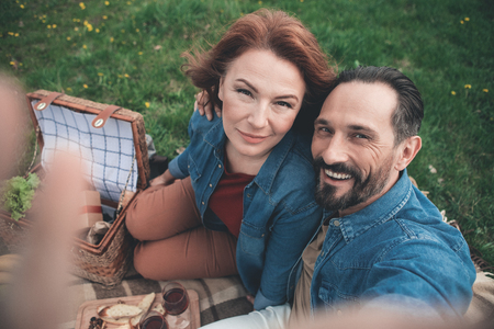 Lets remember every happy moment together. Top view portrait of joyful husband and wife are taking photo of themselves while having picnic in the nature. Man is stretching hand up and embracing womanの写真素材