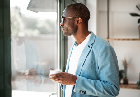 Side view african pensive male drinking cup of delicious coffee while looking at windowの写真素材