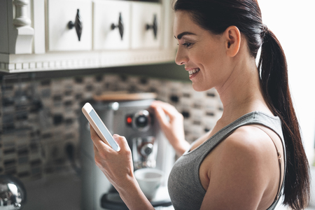 Profile of smiling woman making coffee at home. She is standing by coffee machine and holding mobile. Happy lady is looking at screen with delightの写真素材