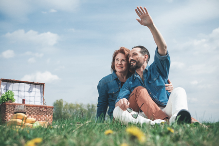 Hello. Cheerful mature man is waving hand to someone while sitting on blanket with his wife. Woman is embracing man and smiling. They are having picnic on the meadowの写真素材