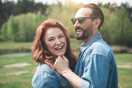Waist up portrait of happy mature loving couple resting in the nature together. They are standing near the river and cuddlingの写真素材