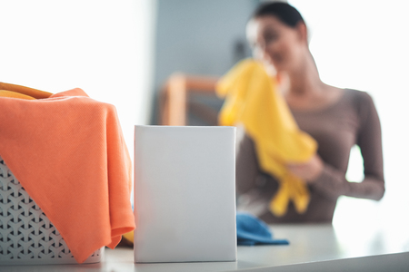 Focus on white packet of washing powder lying on white board. Wicker basket for laundry covered with towel is aside. Woman holding yellow t-shirt is on backgroundの写真素材