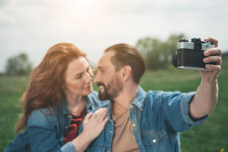 Affectionate married couple is using camera while resting on the grassland. They are looking at each other with love and hugging. Focus on digital deviceの写真素材