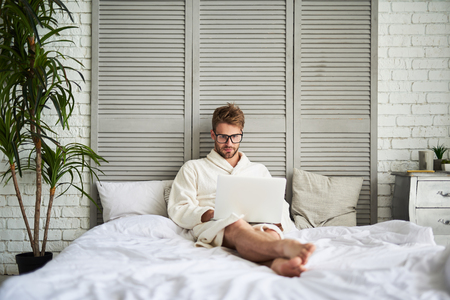 Full length portrait of young man using laptop in bed. He is sitting on white linen wearing bathrobe and eyeglasses. Handsome guy is concentrated on chatting onlineの写真素材