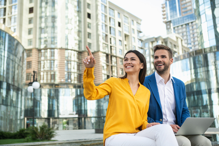 Look there. Cheerful couple is relaxing outdoor in town. Woman is pointing finger up with excitement and laughing. Man is sitting and holding laptopの写真素材
