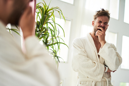 Waist up portrait of drowsy man yawning after night. He is standing in front of mirror in bathrobe and covering face with handの写真素材