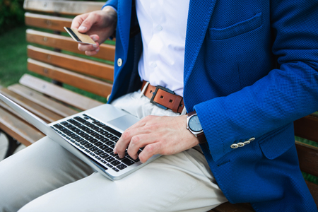 Close up of male arm doing online shopping while holding laptop. Man is sitting on bench outdoorの写真素材