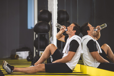 Full length side view pleased man drinking delicious beverage after hard workout in gym. He sitting on floor thereの写真素材