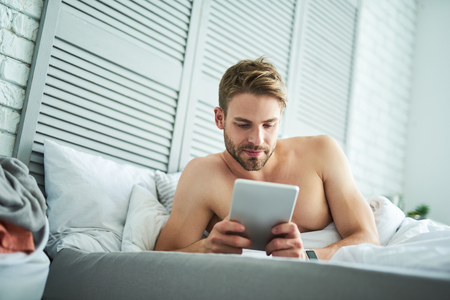 Carefree male lying in bed with tablet in hands. He is watching at screen and enjoying new day at homeの写真素材