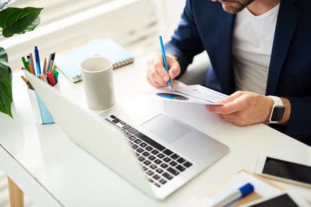 Close up of male hand writing on papers. Man is sitting at table and holding documents with charts while making notes with blue pencil. Laptop and coffee mug are asideの写真素材