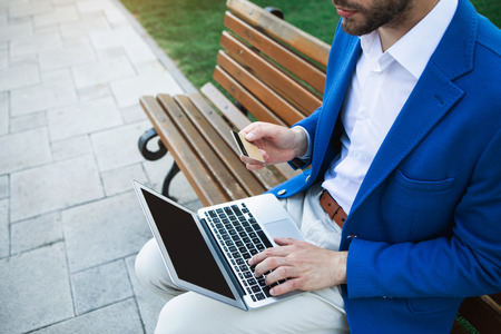 Top view of successful businessman purchasing in internet. He is holding his credit card while typing on laptop. Guy is sitting on bench in parkの写真素材