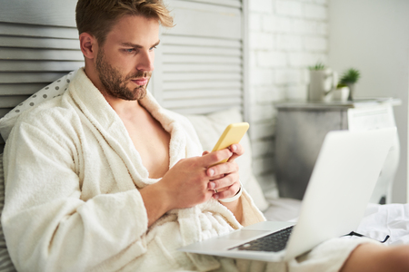 Side view of focused male sitting on bed and holding mobile in hands. He is communicating online while working at laptop lying on his kneesの写真素材