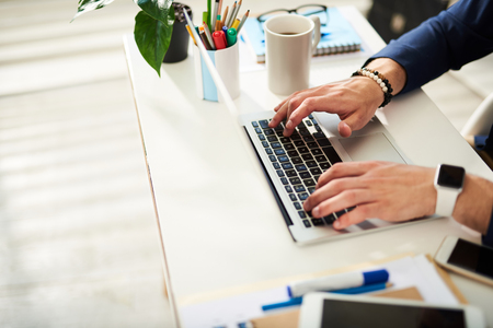 Close up of male hands typing at laptop. Man is sitting and pressing computer keywords. Coffee mug and bunch of colorful markers and pens are asideの写真素材