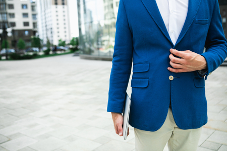 Close up of male elegant suit. Man is buttoning blue jacket while standing on street. He is holding laptop in arm. Copy spaceの写真素材