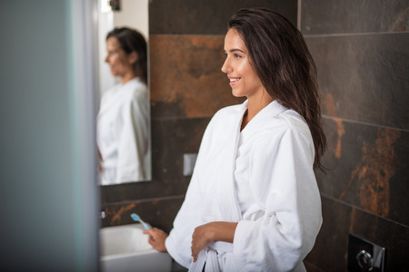 Side view happy female holding toothbrush while locating in bathroom. She reflecting in mirror and...