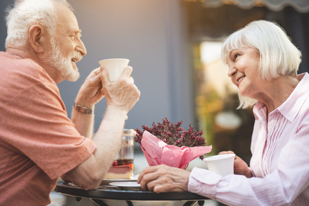 Side view profile of happy senior couple drinking tea in cafe outdoors. They are laughing with delight while having pleasant chatの写真素材