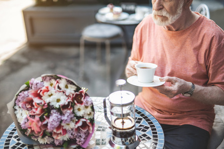 Mature calm male sitting at table with cup of tea and expecting for woman. Bunch of beautiful flowers is lying nearbyの写真素材