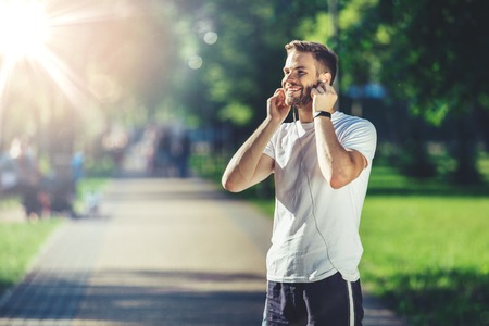 Perfect time for sport. Side view of smiling young man putting earphones in park. He is preparing for jogging outside with delight. Copy space in left sideの写真素材