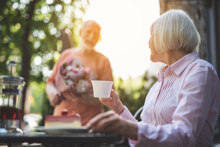 Pleasant expectation. Focus on side view of mature woman sitting at table with cup of tea in hand and turning sideways. Senior male with bunch of flowers is coming over joyfullyの写真素材