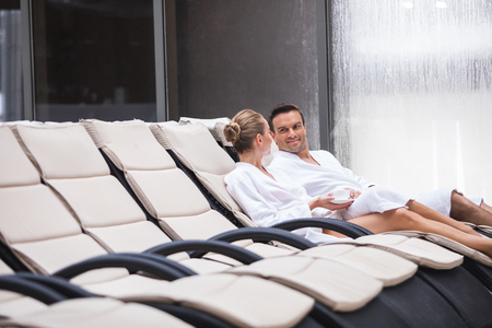 Smiling male and female wearing bathrobes lying on deckchairs. They are drinking coffee and having pleasant talk with delightの写真素材