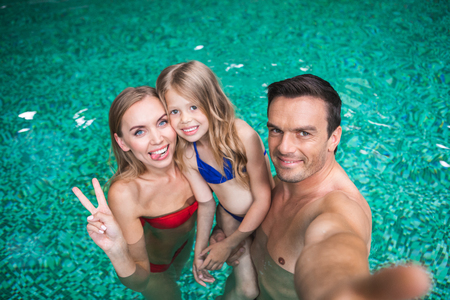 Waist up portrait of smiling parents and child bonding to each other in pool. Woman is showing tongue with joy while man is holding cameraの写真素材