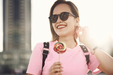 Portrait of happy lady with attractive smile gesticulating arm while eating candy outside. She wearing modern sunglasses during sunny dayの写真素材