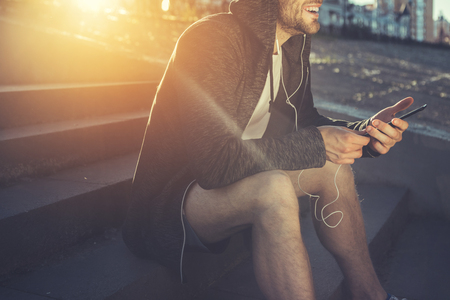 Side view of happy young male sitting on stairs outside and holding smartphone. He is listening to music with earphone and smilingの写真素材
