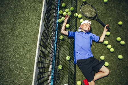Top view portrait of smiling tired child lying on green grass near net around balls for playing tennisの写真素材