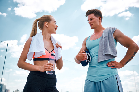 Low angle of cheerful woman and man standing and talking after training in open air. Girl is holding bottle of water while guy is keeping boxing gloves. They are using towels on shoulders and looking at each otherの写真素材
