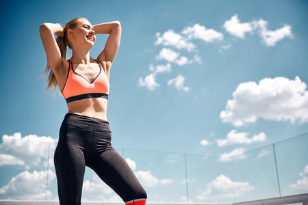 Low angle of carefree sportswoman relaxing and laughing while spending active time. She is standing and putting hands behind head while enjoying sunny weather. Copy space in right sideの写真素材