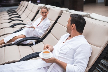 Side view of smiling male and female sitting on deckchairs and drinking tea. Young people are communicating while relaxing with joy and delightの写真素材