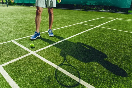 Close up man legs with silhouette standing near ball on green court. He holding racquet in armの写真素材