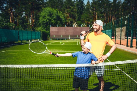 Positive male athlete with cheerful boy playing tennis. Glad kid pointing racket while situating on court near himの写真素材